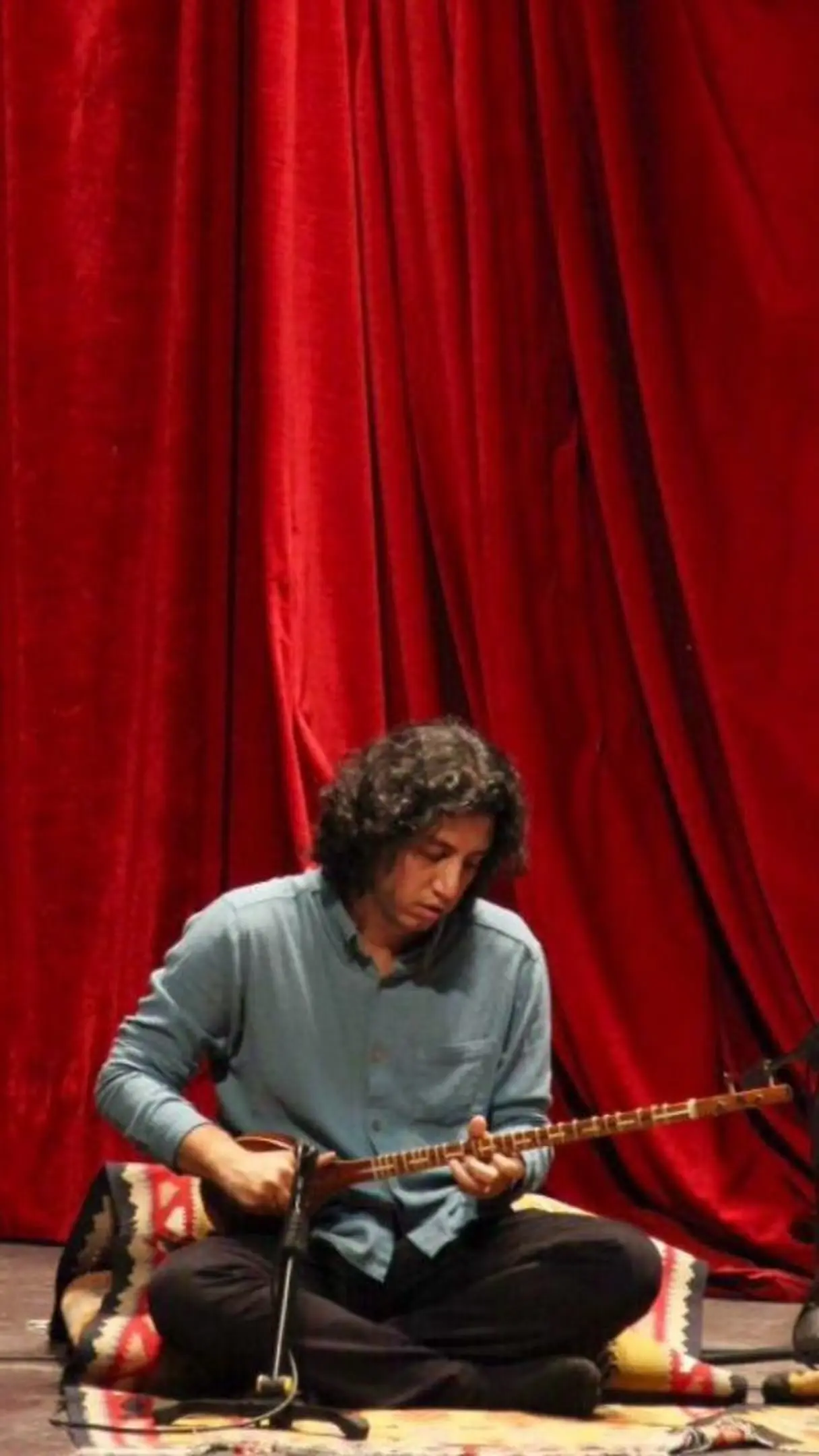 Siavash Karami performing on the setar before a red stage curtain.